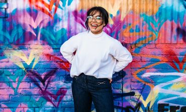 Woman in glasses and white top laughing in front of colourful graffiti wall
