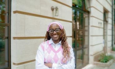 Seun Sowunmi wears a pink hairband, a white shirt, and a pink and white sleeveless jumper. She smiles with arms folded. She wears glasses, and her hair is in braids.