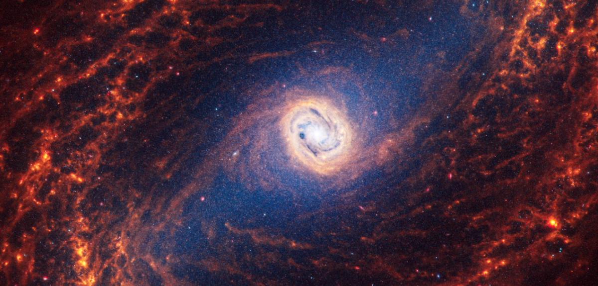Spiral galaxy 1433. The spiral arms are composed of many filaments in shades of orange. Thin dust lanes connect from the core, through the bar to the spiral arms. The galaxy’s arms begin at the end of the bar and appear to rotate counterclockwise. 