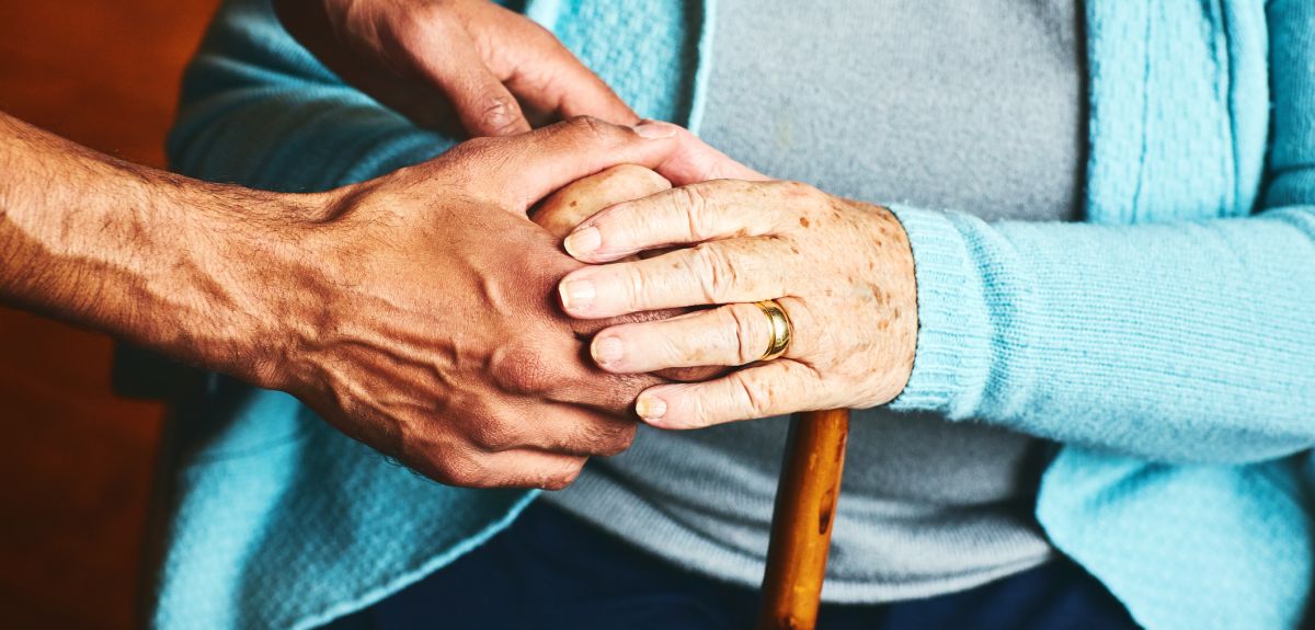 Close up of a carer holding hands of seated senior woman, who wears a blue cardigan and has a walking stick.
