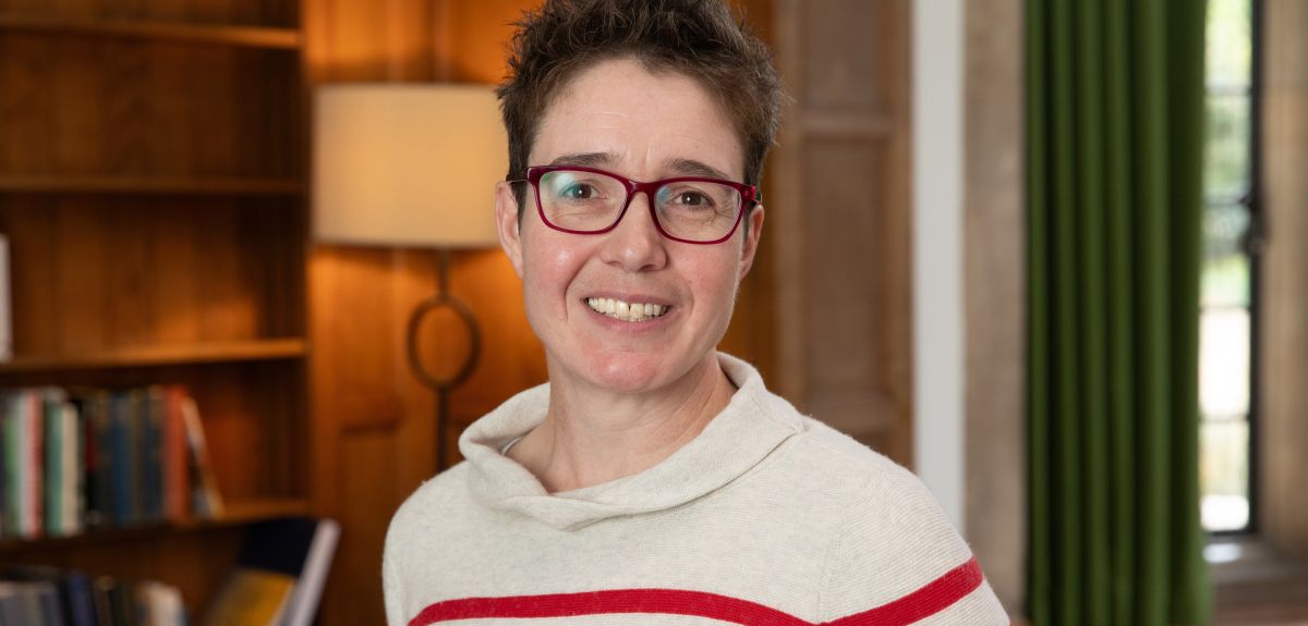 A portrait photo of Charlotte Deane, a white lady with short brown hair wearing glasses and a stiped jumper, standing by a bookcase.