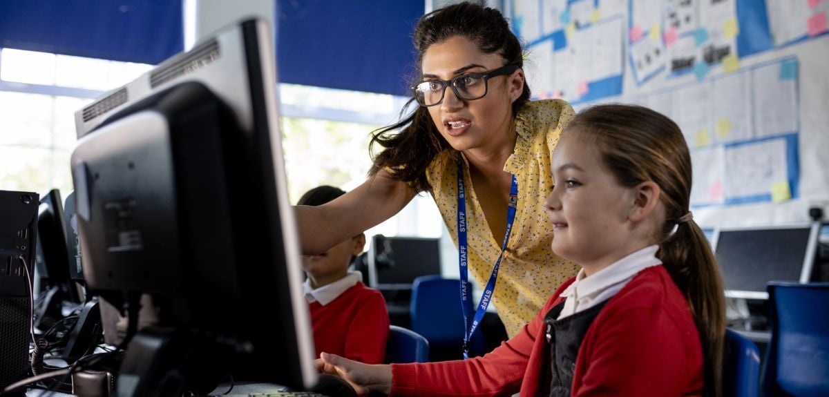 A close-up front view of a teacher and her young pupils who are taking part in a computing class. The teacher is offering one of the girls in her class some advice as she gets to grips with the computer.