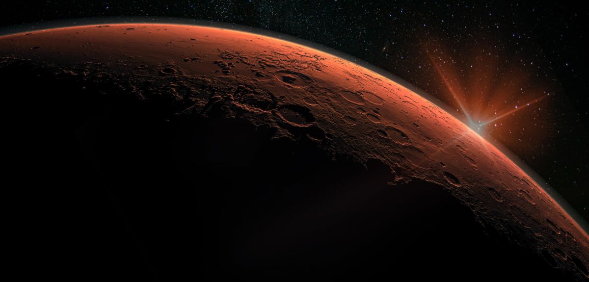 A sunrise as seen from Mars. The red surface of the planet is illuminated by the disc of the sun appearing on the horizon. The background is black outer space with stars. 
