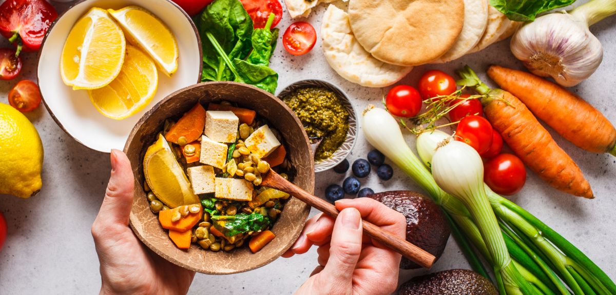 A spread of plant-based foods including flat breads, tomatoes, spring onions, oranges, carrots, blueberries, and garlic. In the foreground, two hands hold a bowl of lentil curry with tofu. 