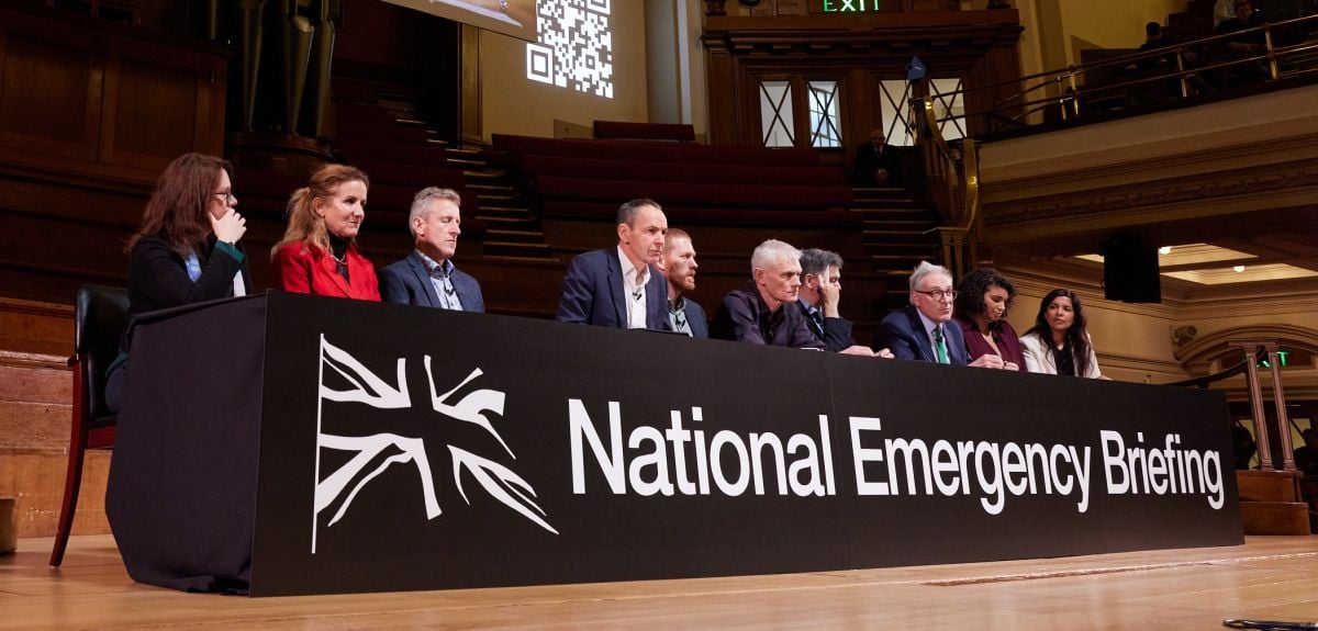 A panel of men and women sit at a table with a table cloth saying 'National Emergency Briefing' and a Union Jack flag.