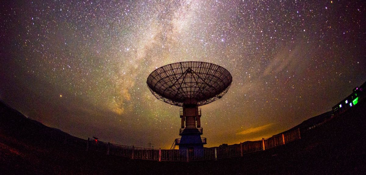 A radio telescope pointing upwards is silhouetted against the Milky Way, seen as a vast array of stars in a twilight sky. 