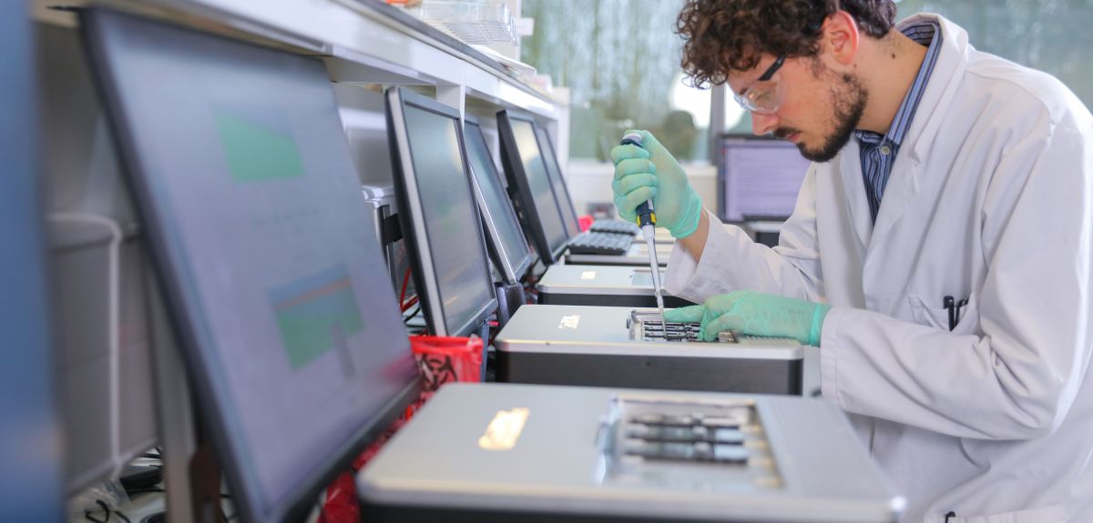 A male scientist in a white coat loads a sample into a machine using a pipette. 