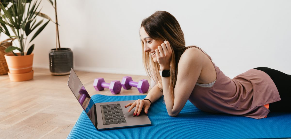 Woman on a yoga mat, looking at a laptop. Purple dumbbells and plants in the background