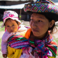 A young Peruvian woman wearing a colourful hat and shawl holds a baby. She is standing outside. Village buildings are visible behind her.