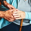 Close up of a carer holding hands of seated senior woman, who wears a blue cardigan and has a walking stick.