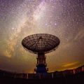 A radio telescope pointing upwards is silhouetted against the Milky Way, seen as a vast array of stars in a twilight sky. 